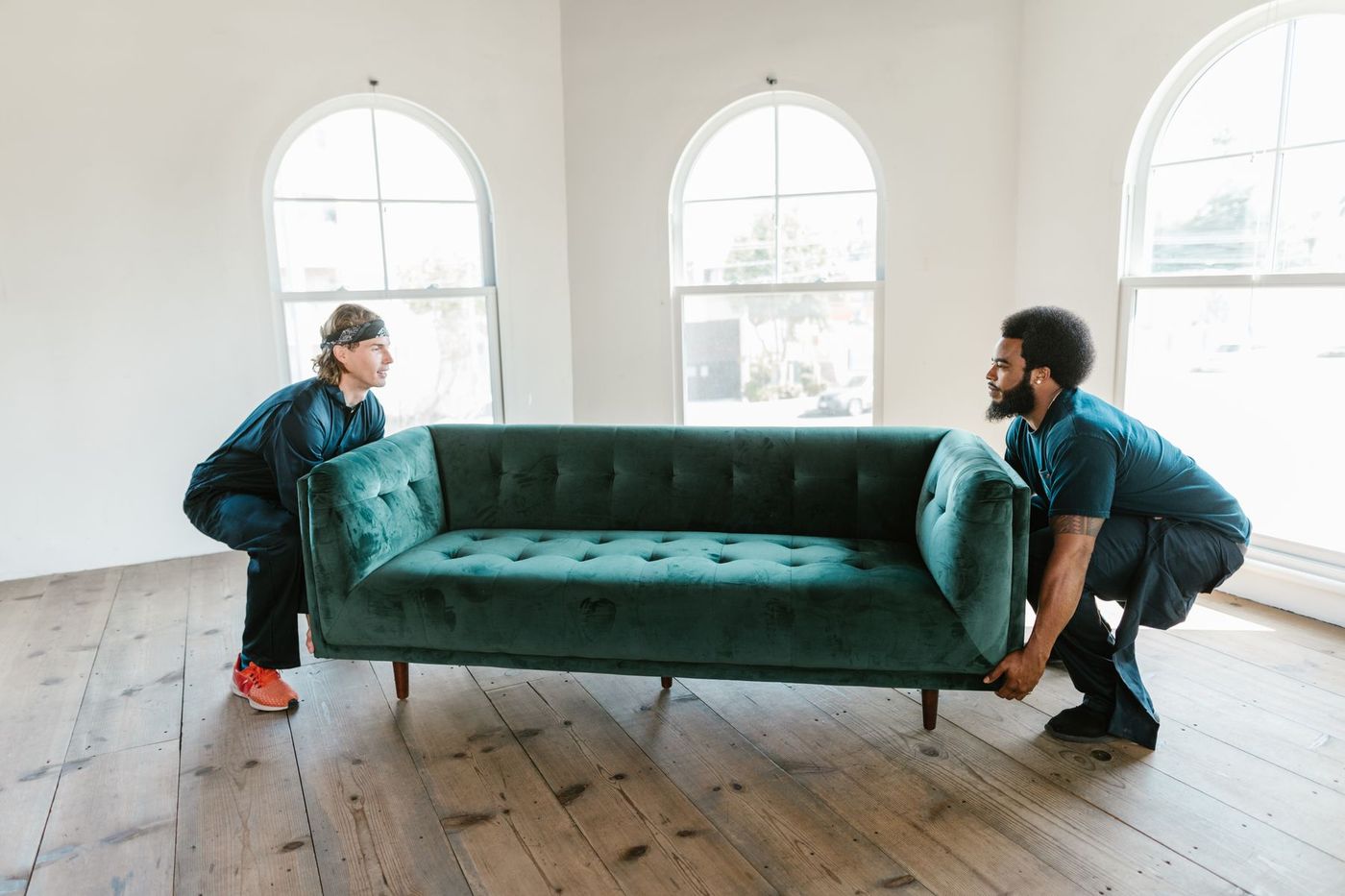Two movers lifting a velvet sofa together in a bright, empty room with tall arched windows