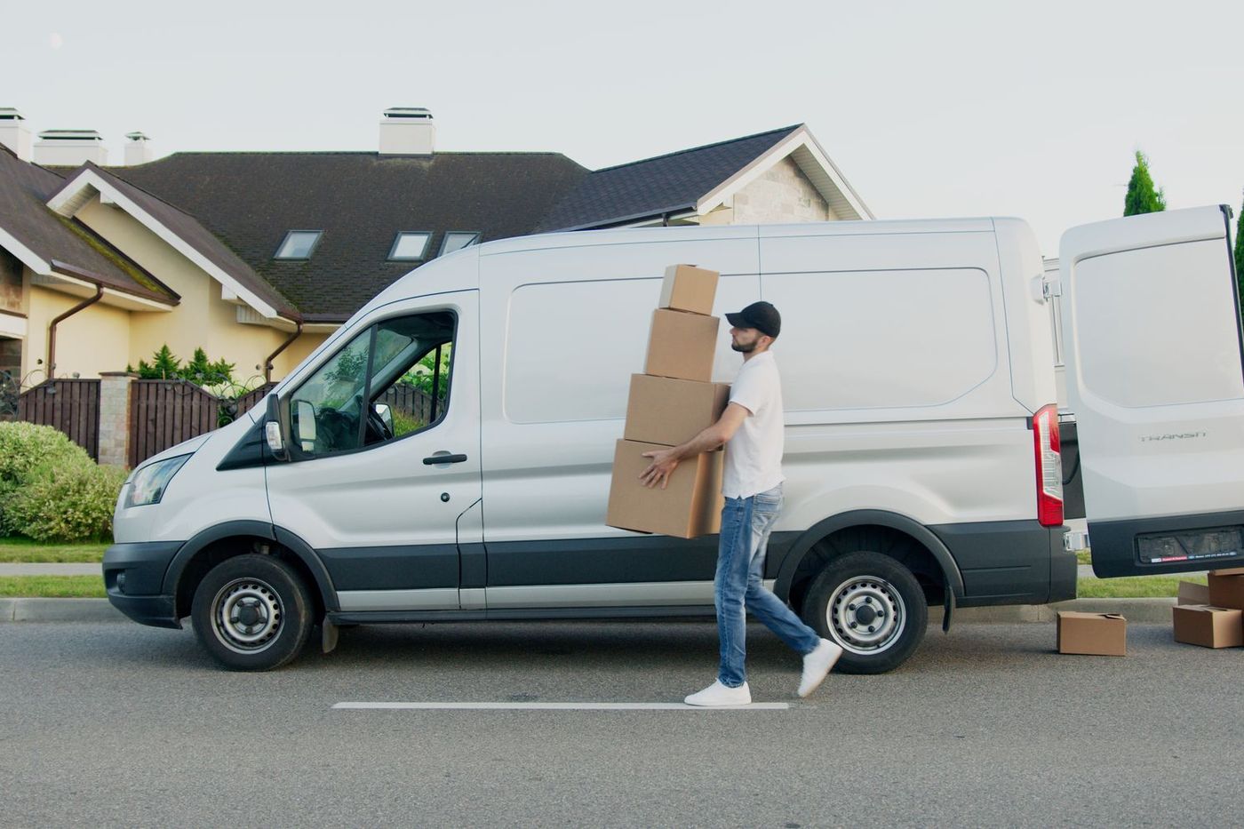 A plain white long-wheelbase van parked at the kerb while a mover carries a stack of cardboard boxes towards it