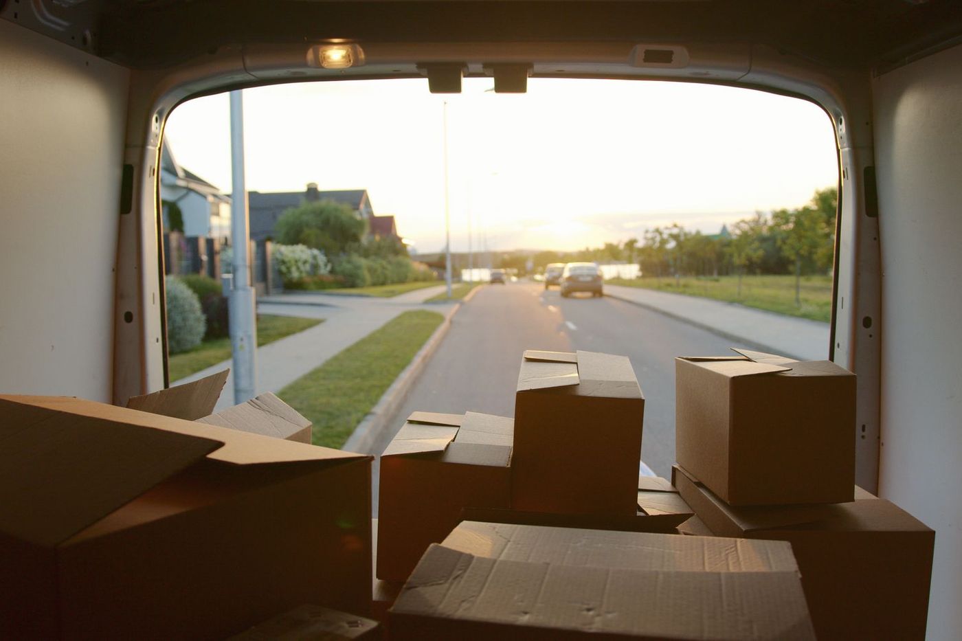 Looking out through the open rear doors of a loaded van onto a suburban street at dusk, boxes stacked inside